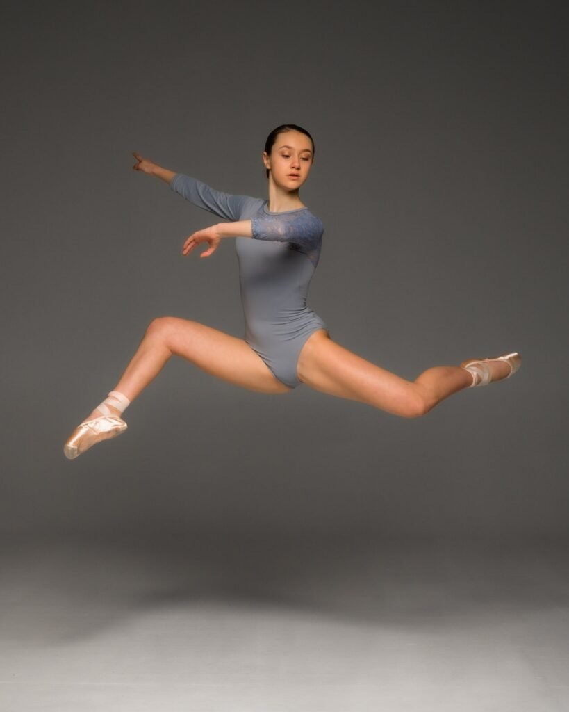 Dancer mid-leap in a dusty blue leotard with lace shoulder